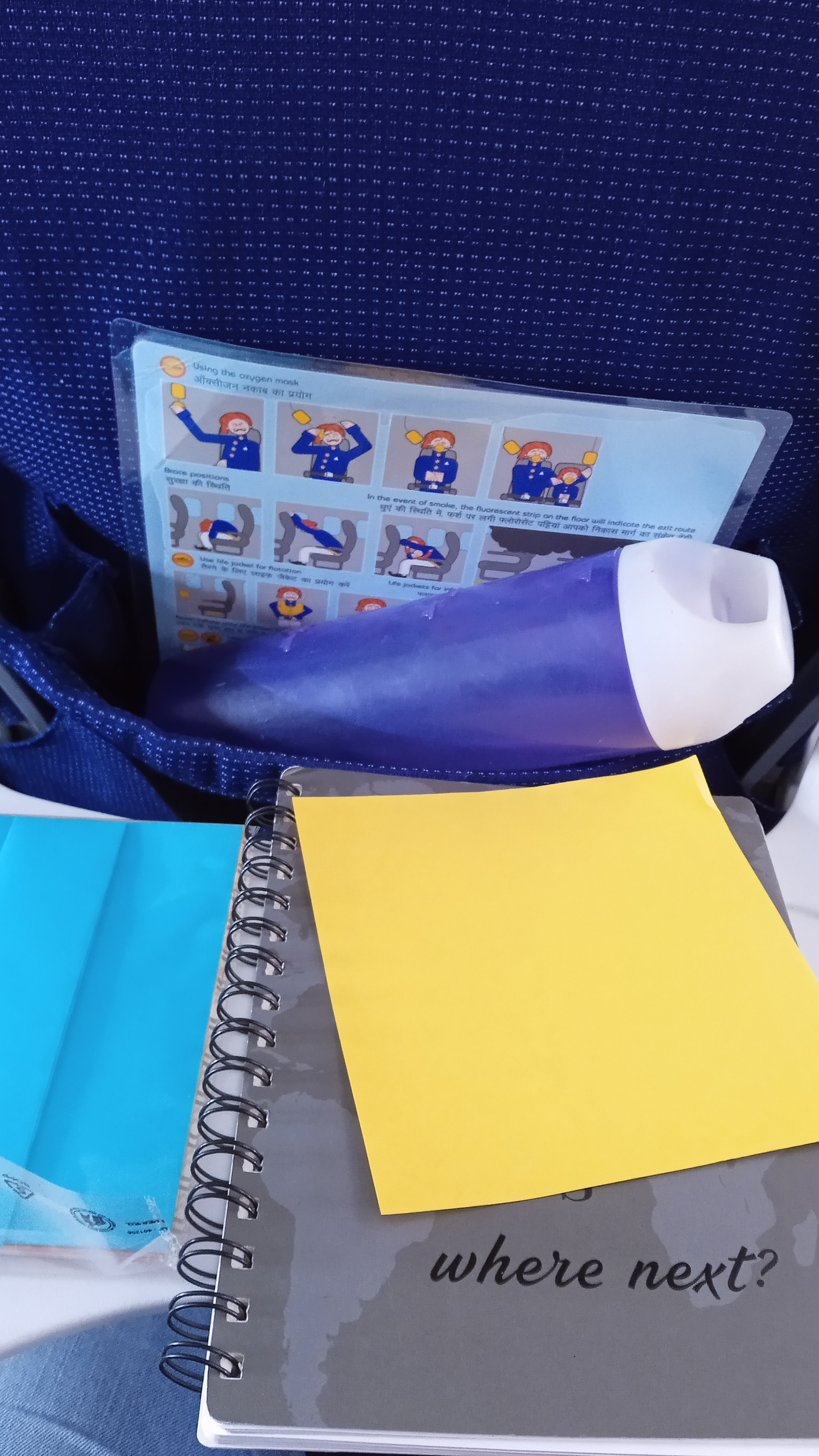 notebook, pen, and kami placed on a flight seat tray, and a water bottle in the seat pouch.